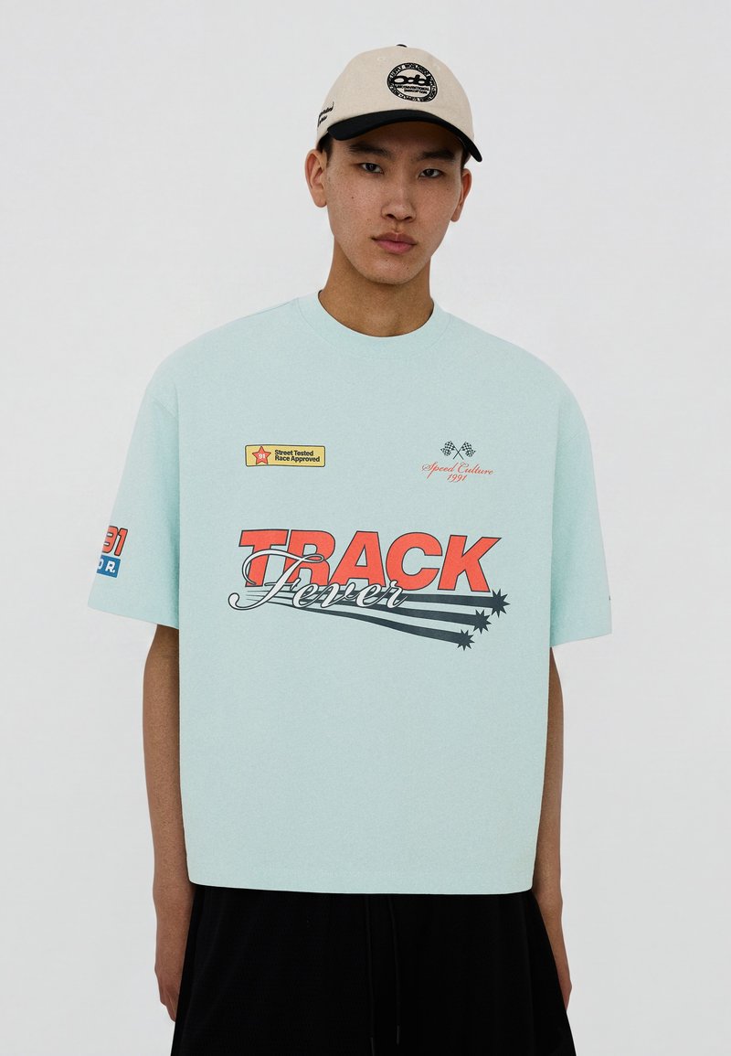 Jeune homme portant un T-shirt oversize bleu clair avec le texte "TRACK Fever" et une casquette de baseball beige sur un fond uni.