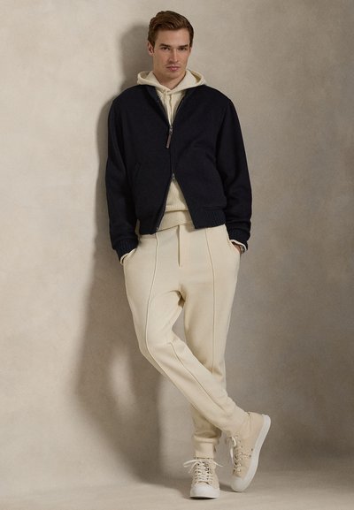 Μπλε ναυτικό μπουφάν bomber πάνω από μια μπεζ φούτερ με κουκούλα, συνδυασμένο με μπεζ παντελόνια και μπεζ αθλητικά παπούτσια. Το μπουφάν έχει ριμπ μανσέτες και φερμουάρ κλείσιμο.