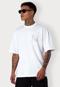 White cotton t-shirt with short sleeves. Features multicolored geometric print on the left chest. Model wears black sunglasses. Black pants.