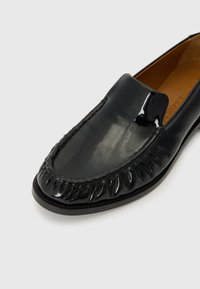 Črne lakirane loferice z zaobljenim nosom, dekorativnimi resicami in kontrastnim šivom ob robu. Gladka tekstura.