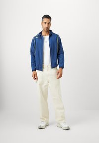 Blå lättviktsjacka med gula detaljer, vit linne, krämfärgade chinos och grå sneakers. Slät textur, avslappnad passform.