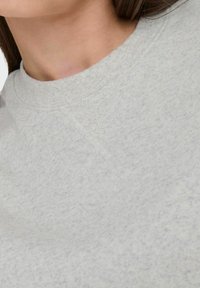 Detailní záběr na osobu v svetru s kulatým výstřihem světle šedé barvy, na kterém je pod límcem vidět vyztužení ve tvaru V se složitým prošíváním.