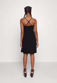 Black sleeveless dress with a fitted bodice and flared skirt, featuring crisscross straps and a smooth texture. Worn with chunky heeled sandals.