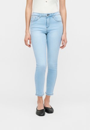 Kvinde iført lyseblå skinny jeans og sorte flade sko, stående mod en ensfarvet hvid baggrund.