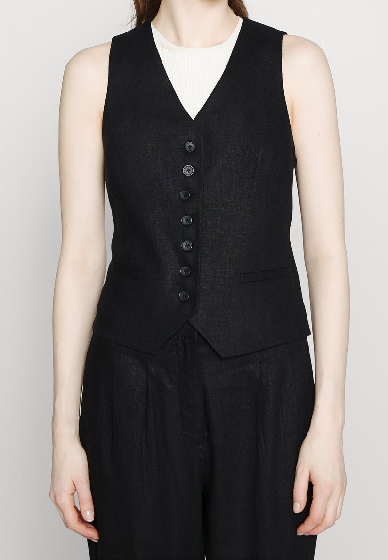 Personne portant un gilet noir boutonné sur un haut blanc, associé à un pantalon noir, montré des épaules aux hanches sur un fond uni.