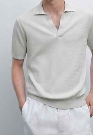 Mann trägt ein hellbeiges kurzärmeliges Poloshirt, in weiße Hose gesteckt, Hände in den Taschen, vor schlichtem hellem Hintergrund.
