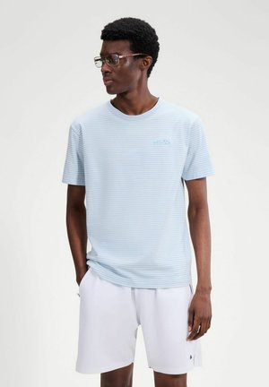 Junger Mann trägt ein hellblau gestreiftes T-Shirt und weiße Shorts, schaut seitlich mit den Händen in den Taschen vor einem einfarbigen Hintergrund.