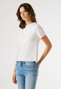 Λευκό κοντομάνικο μπλουζάκι με κυματιστά άκρα, ριμπ ύφασμα και εφαρμοστή γραμμή, συνδυασμένο με ανοιχτόχρωμο τζιν παντελόνι.