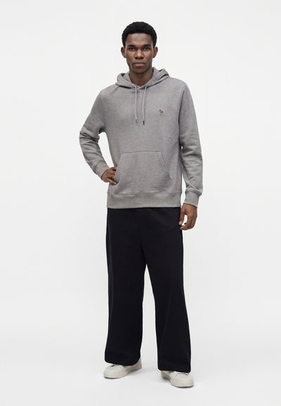 Homme debout, portant un sweat à capuche gris avec poche avant, pantalon large noir et baskets blanches, sur un fond clair uni.