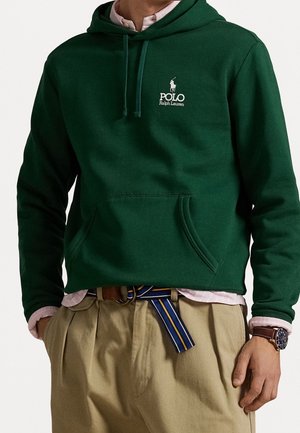 Mann trägt dunkelgrünen Polo Ralph Lauren Hoodie, hellrosa Hemd, beige Hose mit gestreiftem blauem Gürtel und eine Armbanduhr am linken Handgelenk.