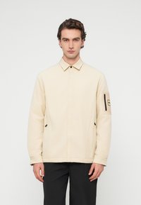 Νέος άντρας που στέκεται με το πρόσωπο προς τα εμπρός, φορώντας ένα μπεζ μπουφάν με τσέπη με φερμουάρ στο αριστερό μανίκι και μαύρο παντελόνι.