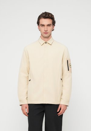 Tânăr care stă cu fața înainte, purtând o jachetă crem cu un buzunar cu fermoar pe mâneca stângă și pantaloni negri.