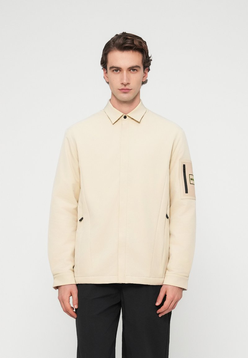 Νέος άντρας που στέκεται με το πρόσωπο προς τα εμπρός, φορώντας ένα μπεζ μπουφάν με τσέπη με φερμουάρ στο αριστερό μανίκι και μαύρο παντελόνι.