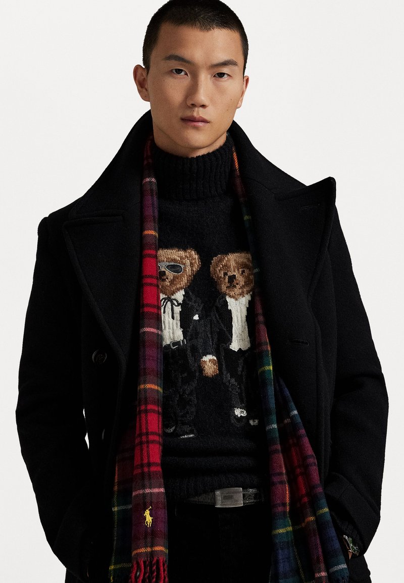 Manteau noir par-dessus un pull col roulé noir avec deux ours en tenue formelle. Écharpe en laine à carreaux avec des accents rouges, verts et jaunes.