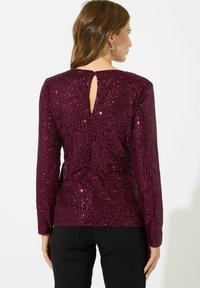 Top bordeaux con paillettes e maniche lunghe, dettaglio a oblò sul retro e texture liscia, abbinato a pantaloni neri.