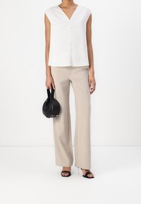 Blouse blanche à col en V profond et manches courtes, associée à un pantalon beige à jambes larges. Porte un petit sac noir, chaussée de talons noirs à enfiler.