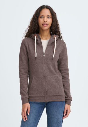 Brun hættetrøje med lynlås, hætte, cremefarvede snørre, forlommer og ribstrikkede manchetter. Bærtes med blå jeans mod en lys baggrund.