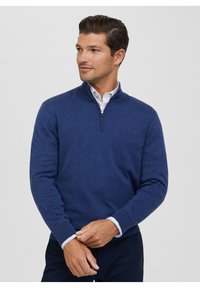 Maglione blu navy con zip e collo alto, realizzato in tessuto morbido, con maniche lunghe e polsini a costine, indossato sopra una camicia con colletto.