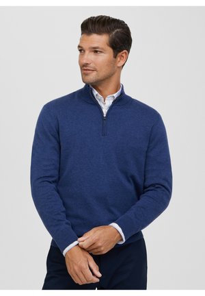 Navy blue zip-neck sweater made of soft fabric, featuring long sleeves and ribbed cuffs, layered over a collared shirt.