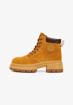 Gelbe Wildleder-Ankle-Boots mit braunem Lederkragen, runder Spitze und klobiger Gummisohle. Mit Metallösen und einem logopatch an der Seite.