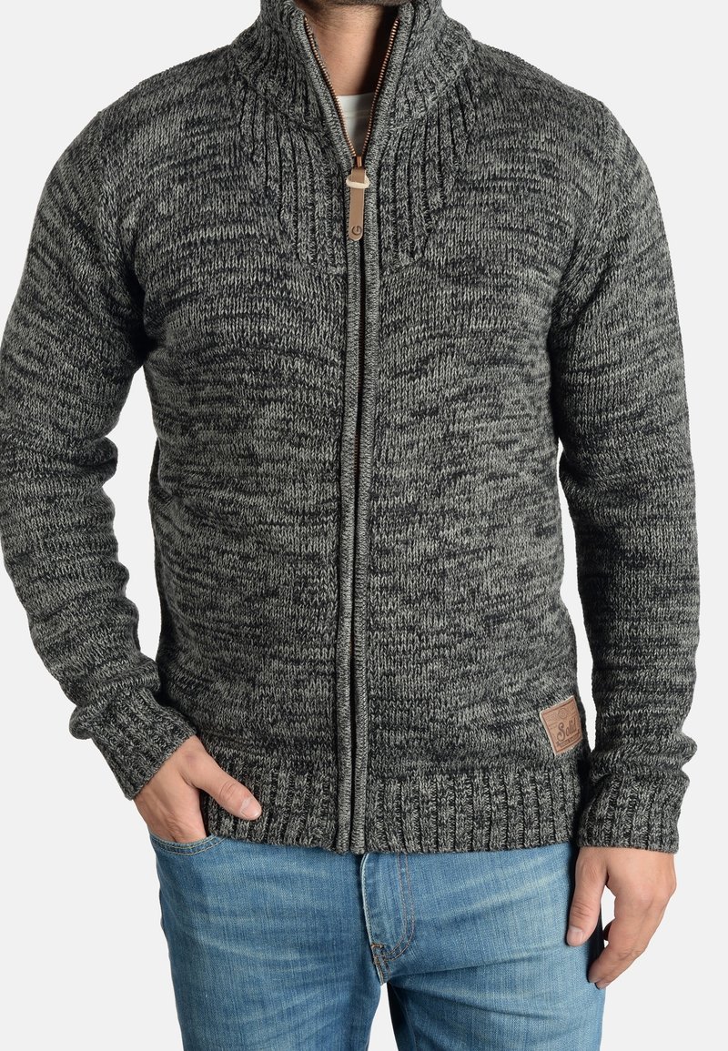 Homme portant un pull en maille gris à fermeture éclair avec un col montant et un jean bleu, une main dans la poche et l'autre sur le côté.