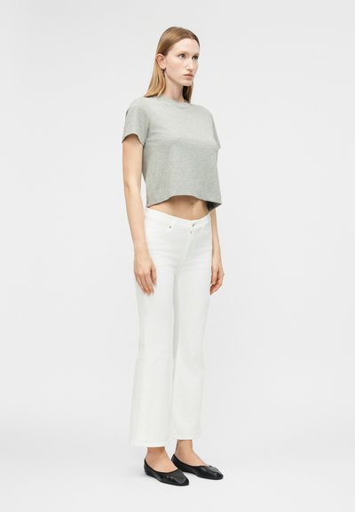 Γυναίκα που στέκεται πλαγίως, φορώντας γκρι κοντομάνικο crop top, λευκό ψηλόμεσο ίσιο παντελόνι και μαύρα φλατ παπούτσια, μπροστά σε απλή φόντο.