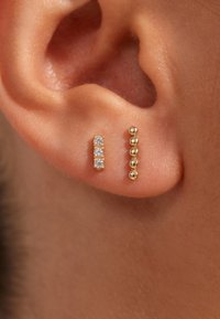 Boucles d'oreilles en or en forme de cerceau avec trois pierres précieuses transparentes et un groupe de petites perles en or, posées sur le lobe d'une personne contre un teint de peau.