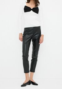 Haut blanc à manches longues avec un détail de nœud noir au niveau du col, associé à un pantalon court en cuir synthétique noir et des chaussures plates noires.