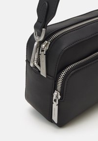 Borsa in pelle nera con due scomparti con cerniera, ferramenta argentata e tracolla rimovibile. Texture liscia con un design minimalista.