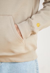 Beige hoodie with a front pocket and ribbed cuffs. Features a yellow embroidered logo on the left sleeve. Soft fabric with a smooth texture.