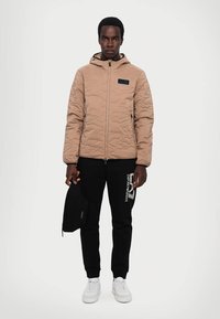 Sandy farvet quiltet jakke med hætte, lynlås foran og sidelommer, parret med sorte sweats med et hvidt logo og hvide sneakers.