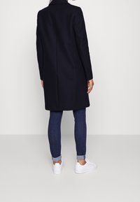 Cappotto di lana blu navy con maniche lunghe, spacco singolo sul retro, indossato sopra jeans scuri e sneakers bianche, con colletto classico.