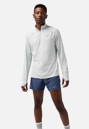 Lys grå langærmet løbetrøje med lynlås krave, parret med navy shorts. Har reflekterende logo og ergonomisk pasform.
