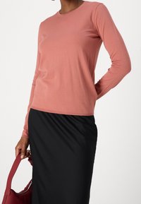 Helles rosa Langarmoberteil aus weichem Strickmaterial, mit Rundhalsausschnitt und lässiger Passform, kombiniert mit einem schwarzen Rock und einer roten Handtasche.