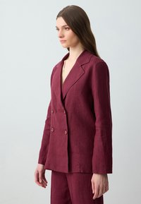 Burgundowa lniana marynarka podwójnie zapięta z klapami i teksturowanym wykończeniem, zestawiona z dopasowanymi spodniami, przedstawiona na jasnym tle.