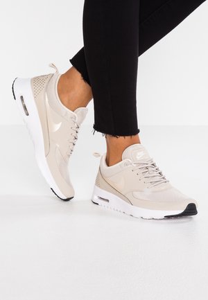 Person iført lyse beige Nike-sneakers med hvide såler og sorte bukser mod en hvid baggrund, stående med den ene fod let løftet.