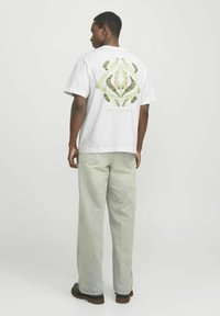 Weißes Baumwoll-T-Shirt mit grünem Blättermuster auf dem Rücken, kombiniert mit hellgrünen, locker sitzenden Jeans und dunklem Schuhwerk.