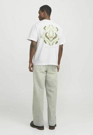 Weißes Baumwoll-T-Shirt mit grünem Blättermuster auf dem Rücken, kombiniert mit hellgrünen, locker sitzenden Jeans und dunklem Schuhwerk.