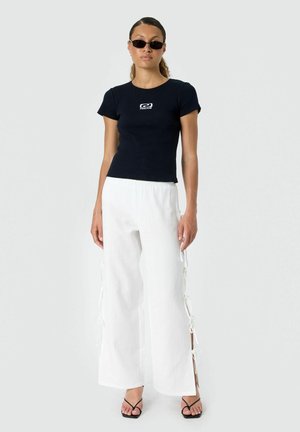 Femme portant un t-shirt noir près du corps, un pantalon blanc à jambes larges avec des liens sur les côtés, des sandales noires à brides et de petites lunettes de soleil noires, debout.