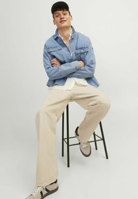 Chaqueta de mezclilla azul claro con bolsillos frontales, camisa blanca debajo, pantalones de pierna ancha beige y zapatillas marrón claro sobre un taburete de metal.