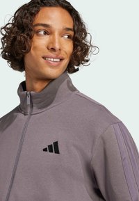 Fialová sportovní bunda s vysokým límcem, zipem a černým vyšívaným logem. Má texturovanou tkaninu a kontrastní pruhy na ramenou.