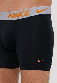 Melnas sporta bokseršortas ar pelēku jostasvietu, uz kuras ir oranžs "NIKE" logo un mazs oranžs "swoosh" logotips apakšējā kreisajā stūrī.