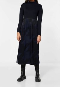 Lång svart klänning med en figurnära överdel och en flödande underdel; har ett hundtandsmönster i mörka nyanser och långa ärmar.