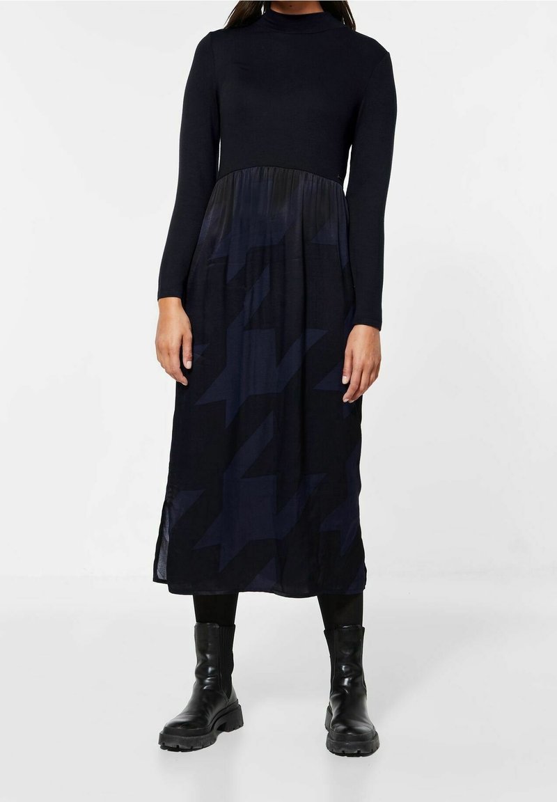 Lång svart klänning med en figurnära överdel och en flödande underdel; har ett hundtandsmönster i mörka nyanser och långa ärmar.