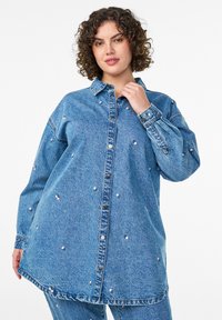 Denim shirt in light blue featuring a button-up front, long sleeves, drop shoulders, and decorative pink flower embellishments throughout.