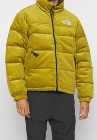 Gul kordflanell-pufferjacka med hög krage, dragkedja och sidofickor. Har justerbara mudd och North Face-logotyp på bröstet.