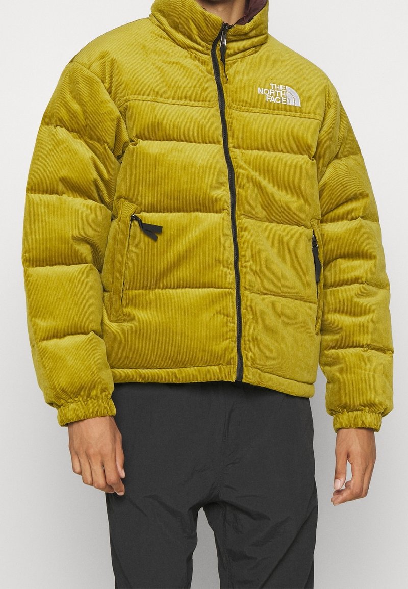 Gul kordflanell-pufferjacka med hög krage, dragkedja och sidofickor. Har justerbara mudd och North Face-logotyp på bröstet.