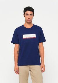 Fiatal férfi sötétkék pólót visel, rajta "JACK&JONES" felirattal, világos bézs nadrággal, egy sima fehér háttér előtt áll.