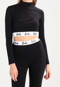 Svart långärmad crop-top med hög halsringning, med ett vitt midjeband med "Barbie"-logotyp. Mjuk, elastisk tyg. Figurformad design.