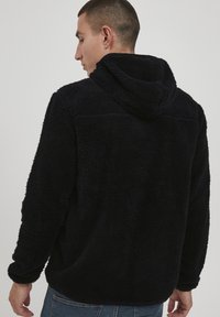 Czarna bluza z polaru o teksturowanej, miękkiej powierzchni, z kapturem i luźnym krojem. Widoczne szwy i casualowy design.